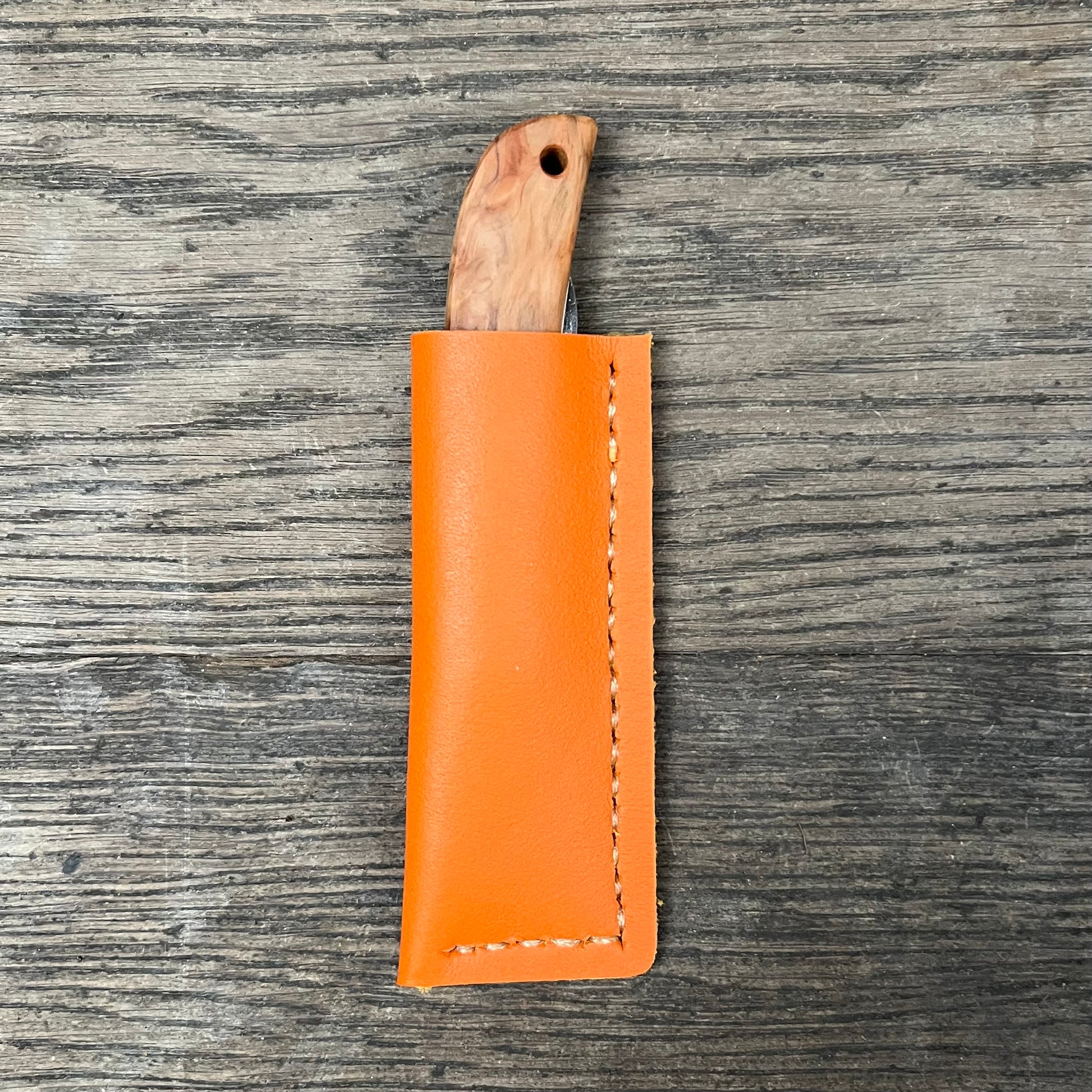 Das 'Kotoh X Niwaki Knife' Taschenmesser von Niwaki mit Holzgriff liegt teils in seiner orangefarbenen Lederschutzhülle auf Holz - ideal für Gartenarbeit oder als zuverlässiges Alltagsmesser.