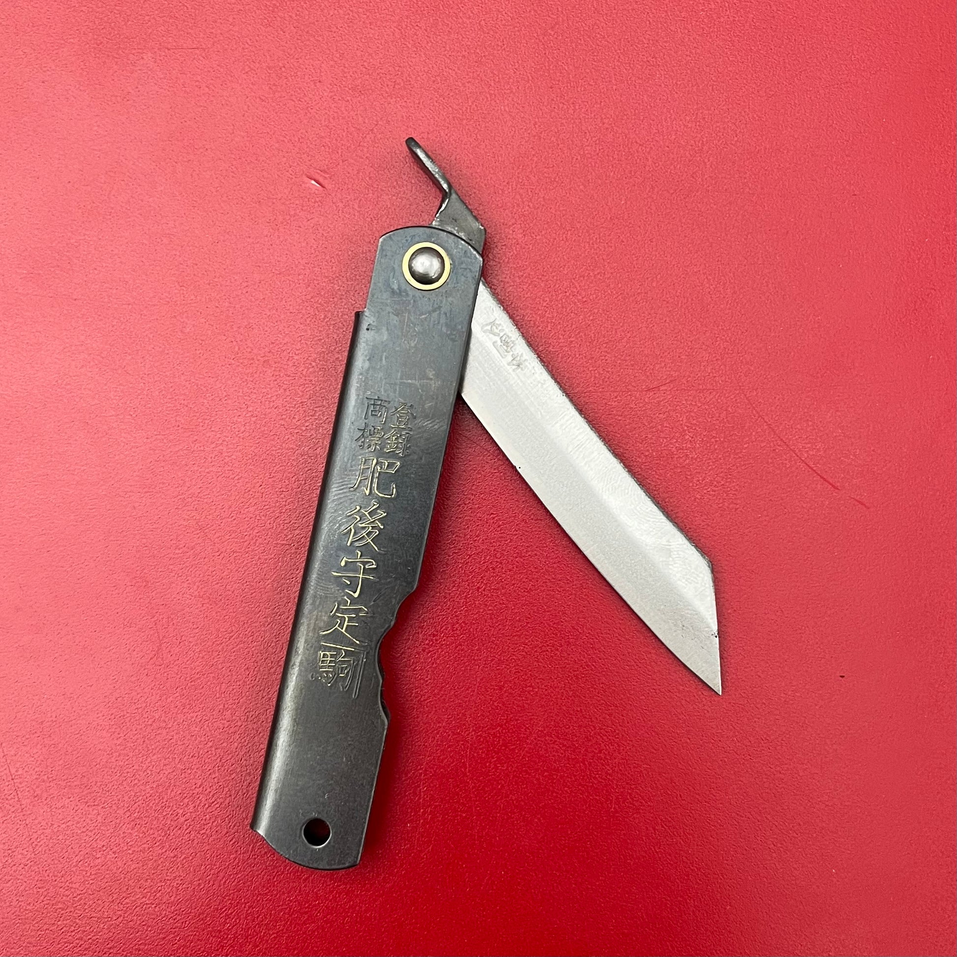 Das Niwaki | 'SK Higonokami Folding Knife' Klappmesser von Niwaki verfügt über eine geradlinige Klinge und einen schwarzen Griff mit japanischer Gravur, der teilweise geöffnet auf einer roten Fläche dargestellt ist.