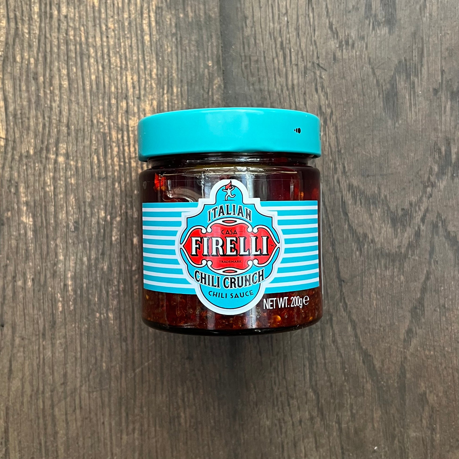 Ein Glas Firelli | Chili Crunch 200g Glas von Firelli, hergestellt mit kalabrischen Chiliflocken und italienischem Olivenöl, steht auf einer dunklen Holzoberfläche. Auf dem türkisfarbenen Deckel und dem Etikett sind der Produktname und das Nettogewicht angegeben.