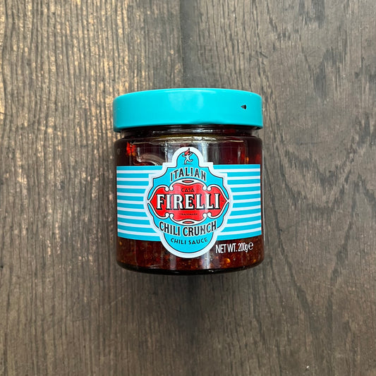 Ein Glas Firelli | Chili Crunch 200g Glas von Firelli, hergestellt mit kalabrischen Chiliflocken und italienischem Olivenöl, steht auf einer dunklen Holzoberfläche. Auf dem türkisfarbenen Deckel und dem Etikett sind der Produktname und das Nettogewicht angegeben.