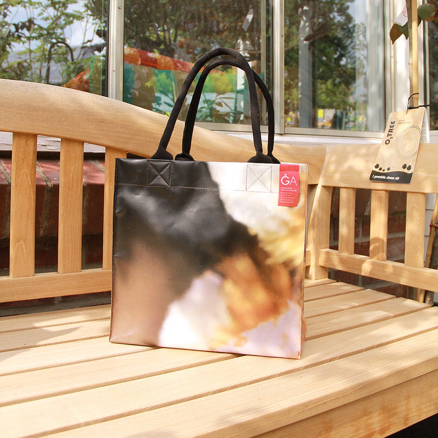 Die Upcycling Tasche 'Torf' aus Werbeplane von Königliche Gartenakademie hat schwarze Griffe und ein unscharfes, abstraktes Design, das im Außenbereich der Königlichen Gartenakademie mit Grünpflanzen und einem Glasgebäude im Hintergrund gezeigt wird.