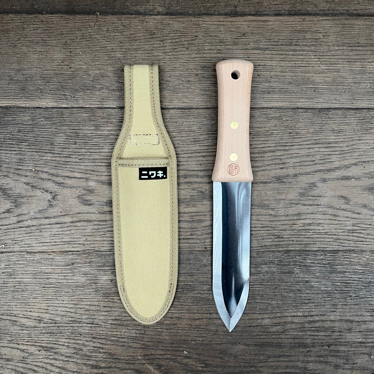 Das Niwaki | 'Hori Hori' multifunktionale Pflanzmesser mit Schutzhülle von Niwaki liegt einsatzbereit auf einer Holzoberfläche - perfekt für Ihre Gartenarbeit.