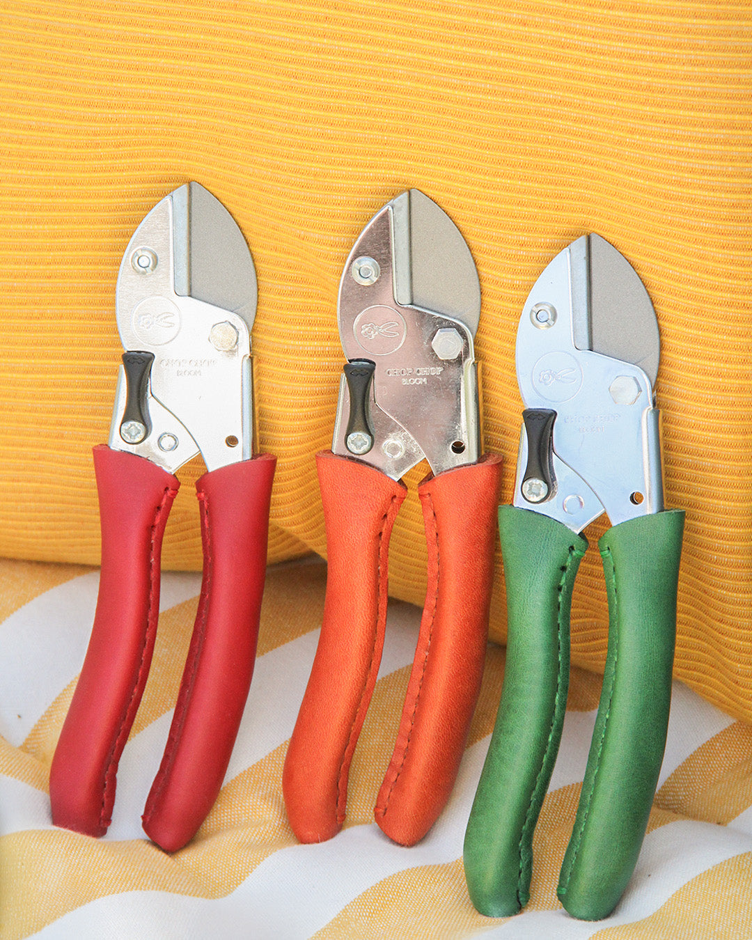 Die Chop Chop Bloom exklusive rote Gartenschere 'Chili Pepper' steht aufrecht vor einem gelb-weiß gestreiften Stoffhintergrund. Sie bringt lebendige Farben und einen Stil, der an eine leuchtende Gartenwerkzeugsammlung erinnert.