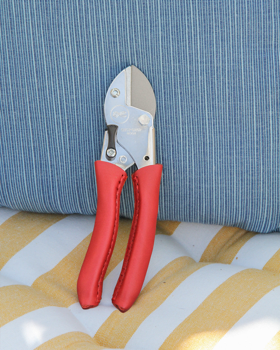 Die Chop Chop | Exklusive rote Gartenschere 'Chili Pepper' von Chop Chop Bloom mit kräftigen roten Griffen sitzt auf blau-weiß und gelb-weiß gestreiften Stoffen.