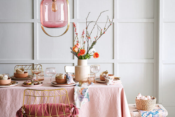Decken Sie einen Osterfest-Tisch mit rosafarbenem Geschirr, goldenen Stühlen, Blumenservietten und einer Vase mit Frühlingsblumen unter einer rosafarbenen Glasleuchte - finden Sie mehr verspielte Deko-Ideen Frühling im "Frühlingstraum" des Callwey Verlags für Ihre festliche Inspiration.