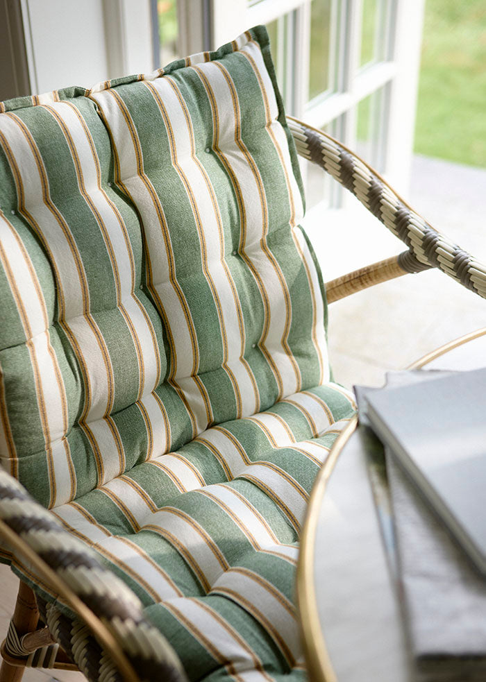 Eine Nahaufnahme des Bungalow 'Palermo Basil' Sitzkissen 45 x 90 cm mit grünen, weißen und beigen Streifen auf einem gepolsterten Stuhl an einem sonnenbeschienenen Fenster, neben einem Tisch mit gestapelten Büchern.