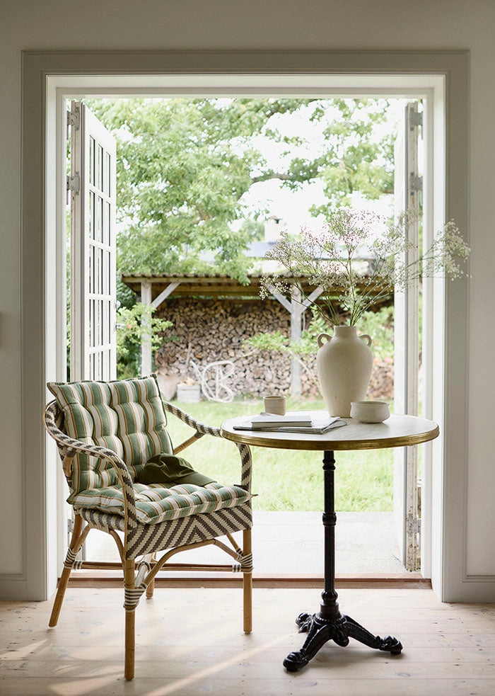 Eine gemütliche Innenraumszene mit einer offenen Tür zu einem grünen Hof. Ein Bungalow 'Palermo Basil' Sitzkissen (45 x 90 cm) liegt auf einem Stuhl neben einem kleinen runden Tisch mit einer weißen Vase mit Wildblumen, einer Tasse und einem Buch; draußen ist gestapeltes Brennholz zu sehen.