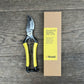 Eine Gartenschere mit gelbem Griff von Niwaki | 'Higurashi GR Secateurs' liegt neben einer braunen Niwaki-Schachtel mit der Aufschrift "Higurashi GR Secateurs" auf Holz; die Gartenschere hat eine silberne Klinge und schwarze Akzente.