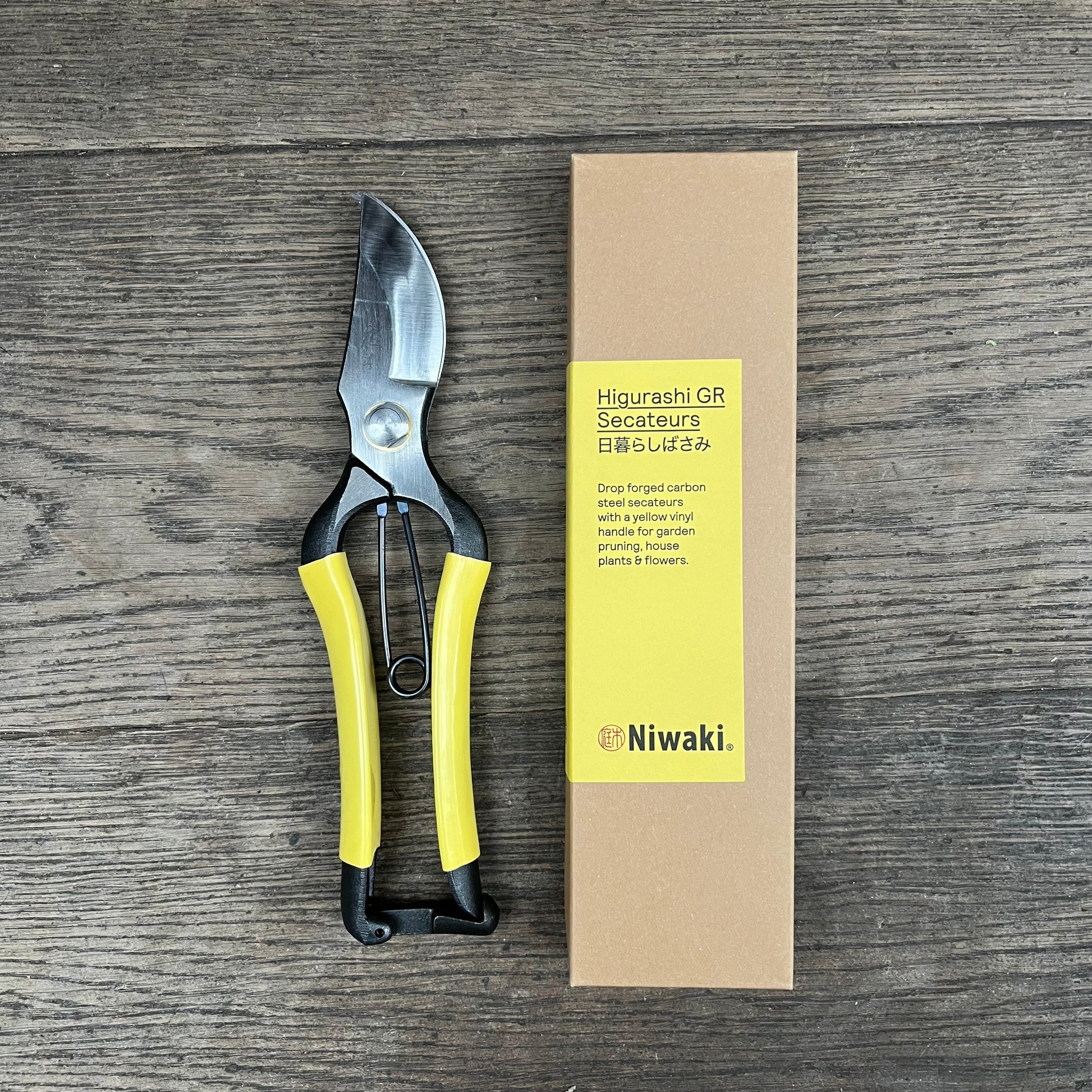 Eine Gartenschere mit gelbem Griff von Niwaki | 'Higurashi GR Secateurs' liegt neben einer braunen Niwaki-Schachtel mit der Aufschrift "Higurashi GR Secateurs" auf Holz; die Gartenschere hat eine silberne Klinge und schwarze Akzente.