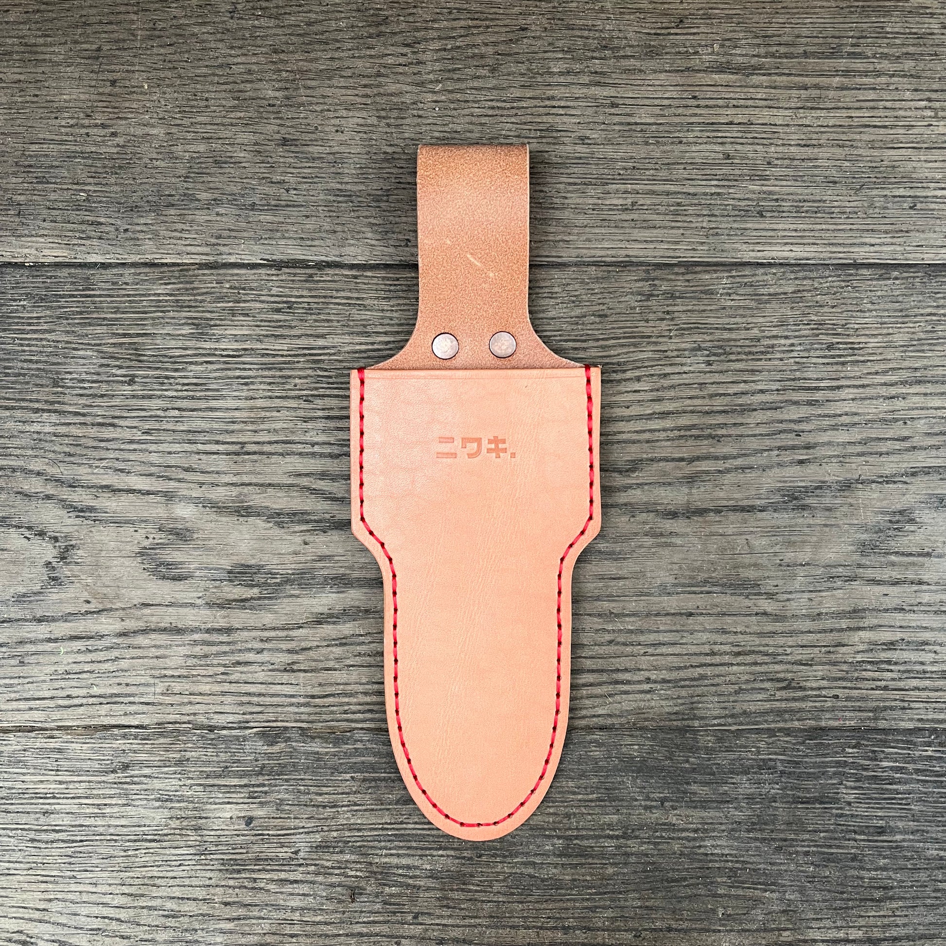 Eine hellbraune Niwaki 'Single Holster' Lederschutzhülle für Schere mit roten Nähten, Metallnieten, geprägten japanischen Schriftzeichen und einer Gürtelschlaufe ruht auf Holz - ideal von Niwaki zum Schutz Ihrer Gartenschere.