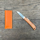 Das Niwaki 'Kotoh X Niwaki Knife' Taschenmesser mit Schutzhülle aus Leder liegt offen auf einer rustikalen Holzoberfläche - ideal für Gartenarbeit oder den täglichen Gebrauch als vielseitiges Taschenmesser.