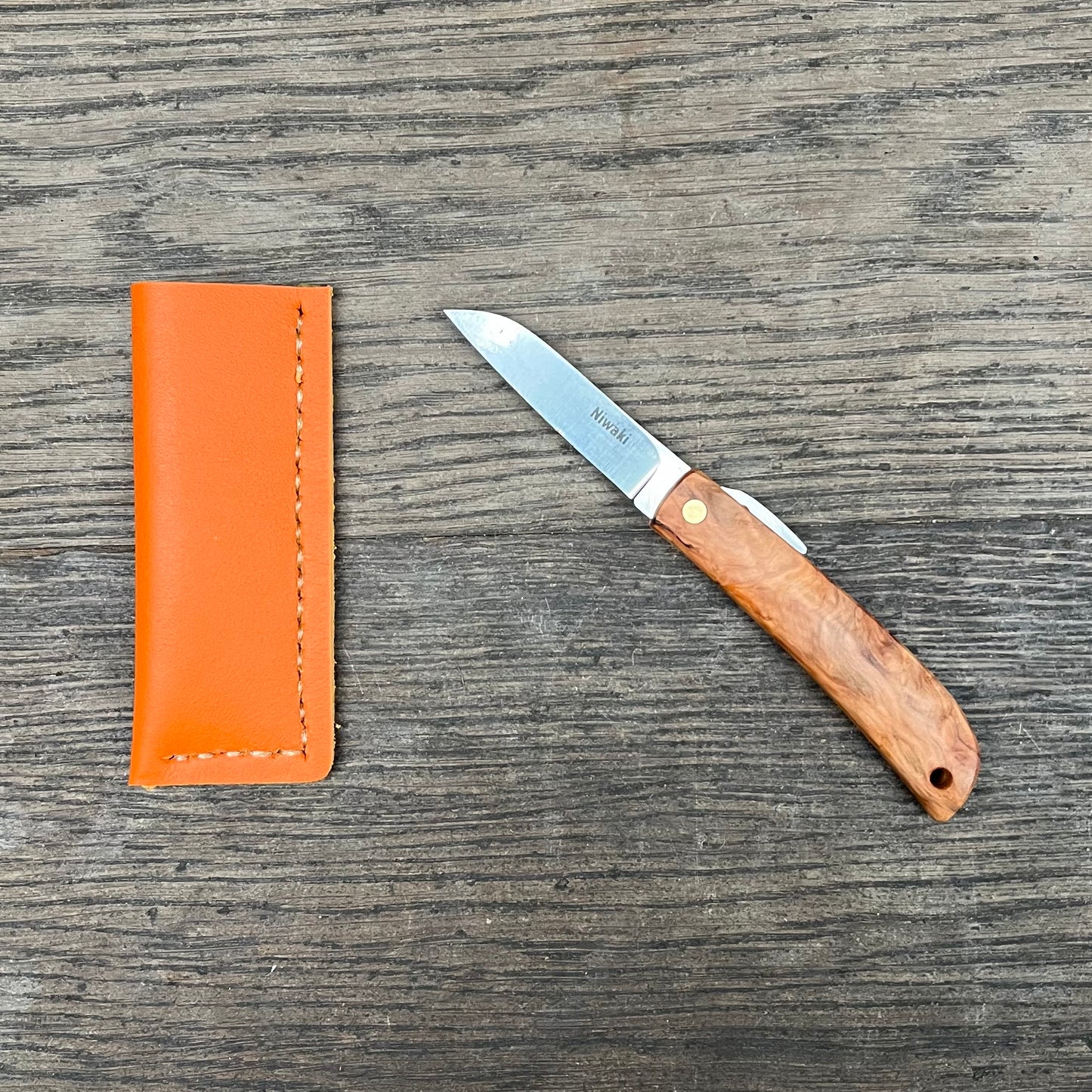 Das Niwaki 'Kotoh X Niwaki Knife' Taschenmesser mit Schutzhülle aus Leder liegt offen auf einer rustikalen Holzoberfläche - ideal für Gartenarbeit oder den täglichen Gebrauch als vielseitiges Taschenmesser.