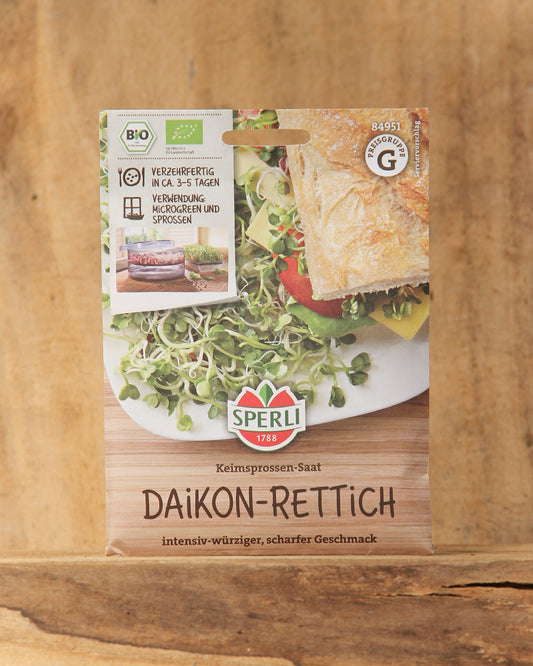 Eine Packung Sperli's Bio Keimsprossen Saatgut 'Daikon-Rettich' wird auf einem hölzernen Hintergrund mit Bildern von frischen Sprossen, Sandwiches und deutschen Produktdetails präsentiert.