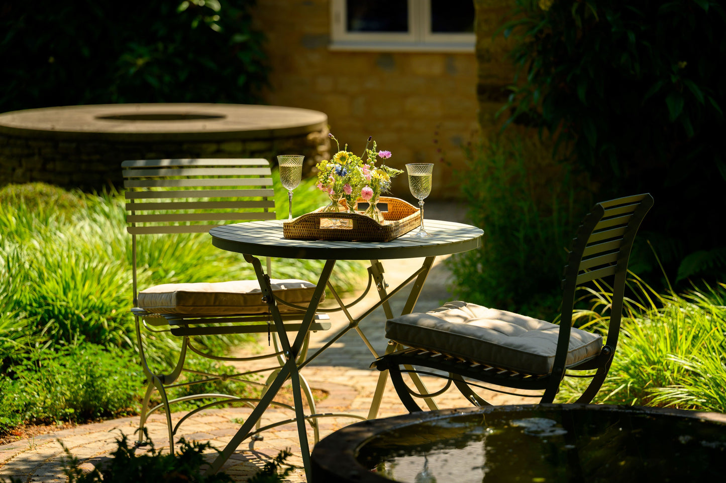 Ein sonnenbeschienener Gartentisch mit dem Harrod Horticultural Bistro Set 'Darsham' - einem klappbaren Gartentisch mit 2 Salbeigrün-Stühlen - mit einem Blumentablett und zwei Gläsern, umgeben von Grünzeug und einem Steinbrunnen im Hintergrund.
