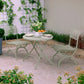 Auf einer kleinen Terrasse im Freien steht das Harrod Horticultural Bistro Set 'Darsham' mit einem klappbaren Gartentisch und 2 Salbeigrün-Stühlen, umrahmt von Grün und Blumen, die an Spalieren entlang einer weißen Backsteinmauer klettern und für eine ruhige Atmosphäre sorgen.
