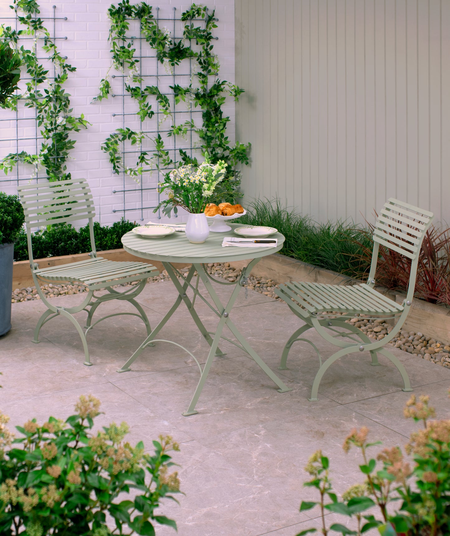 Auf einer kleinen Terrasse im Freien steht das Harrod Horticultural Bistro Set 'Darsham' mit einem klappbaren Gartentisch und 2 Salbeigrün-Stühlen, umrahmt von Grün und Blumen, die an Spalieren entlang einer weißen Backsteinmauer klettern und für eine ruhige Atmosphäre sorgen.