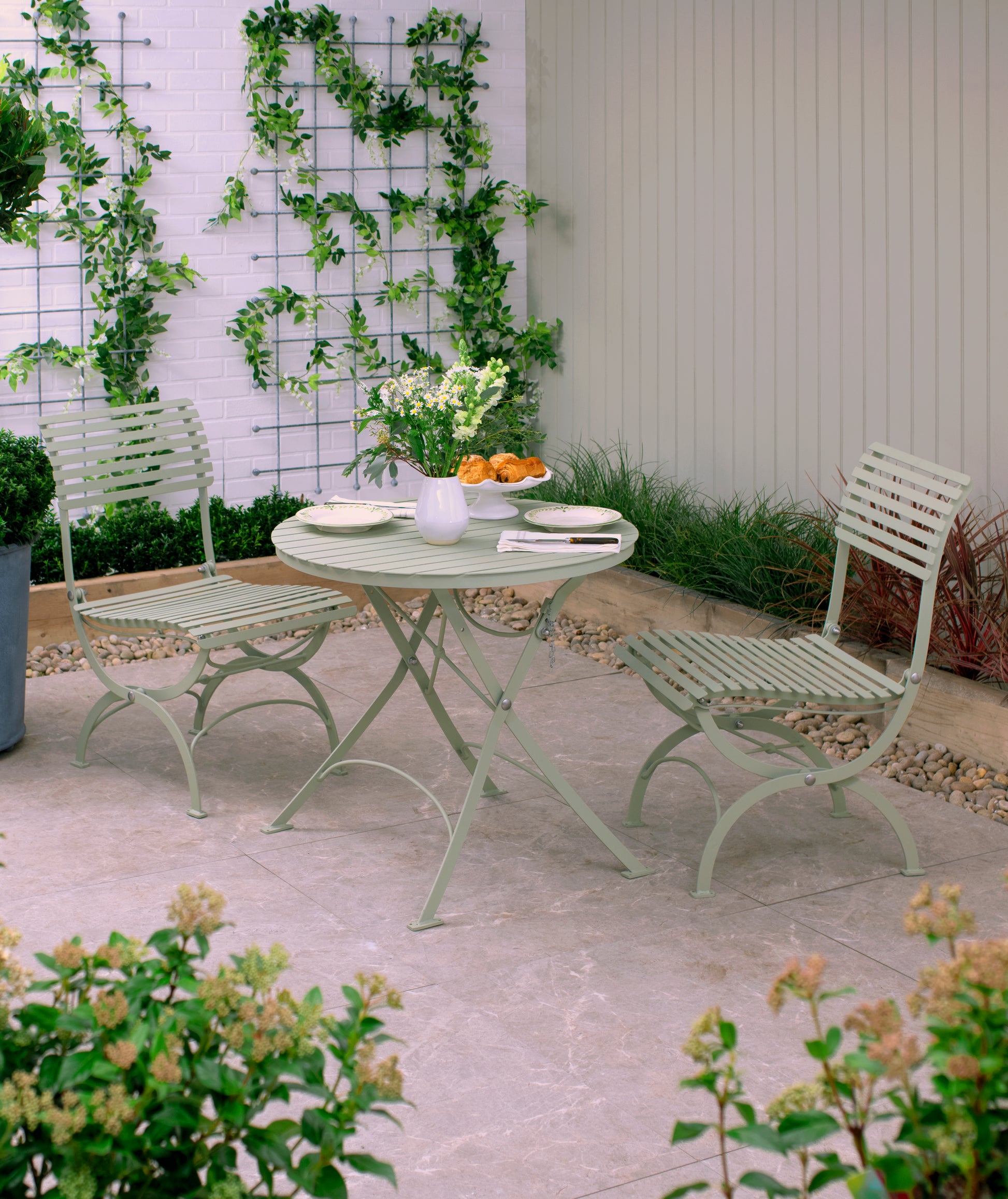 Auf einer kleinen Terrasse im Freien steht das Harrod Horticultural Bistro Set 'Darsham' mit einem klappbaren Gartentisch und 2 Salbeigrün-Stühlen, umrahmt von Grün und Blumen, die an Spalieren entlang einer weißen Backsteinmauer klettern und für eine ruhige Atmosphäre sorgen.