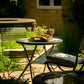 In einem sonnenbeschienenen Garten steht das Harrod Horticultural Darsham Klappbarer Bistro Set mit zwei Stühlen. Auf dem handgefertigten Tisch steht ein Tablett mit einer Blumenvase und zwei weißen Weingläsern, umgeben von üppigem Grün und Steinakzenten.