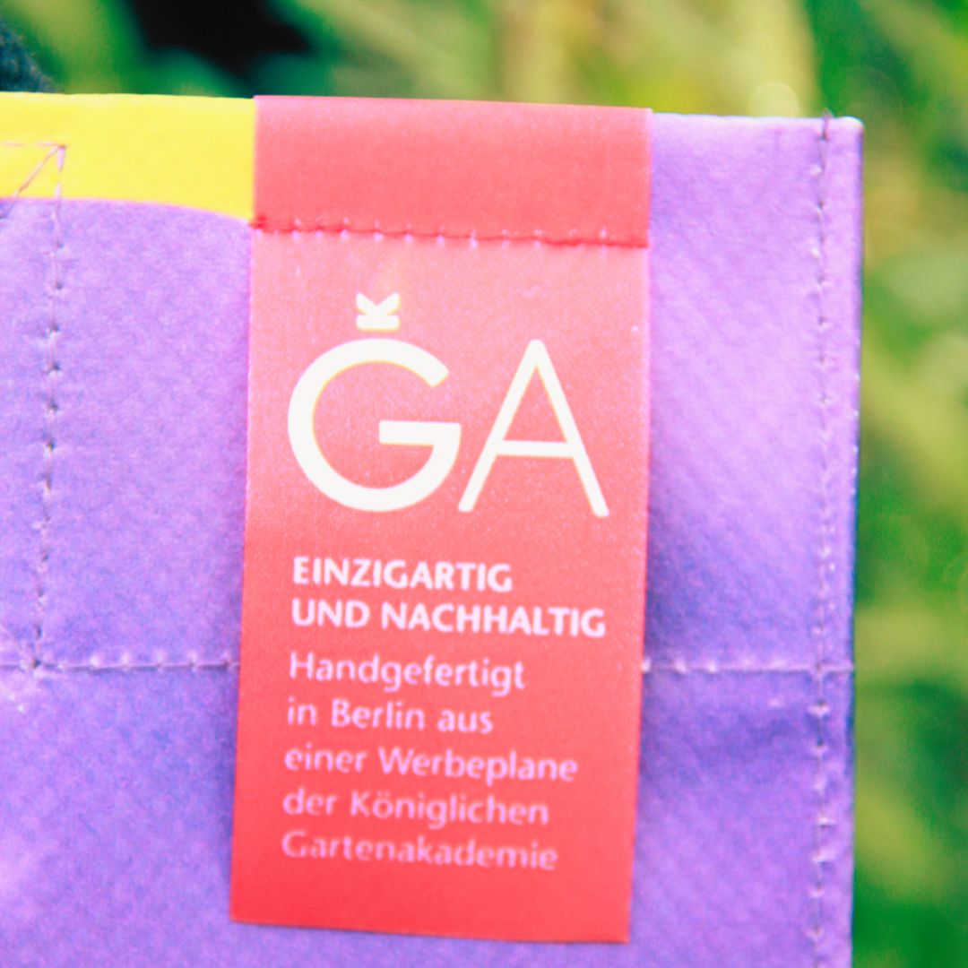 Nahaufnahme der Upcycling KGA Shopper 'Herbs' von Königliche Gartenakademie: aus lila Stoff mit rotem GA-Tag, handgefertigt in Berlin aus einer Werbeplane, einzigartig und nachhaltig. Unscharfer grüner Hintergrund.
