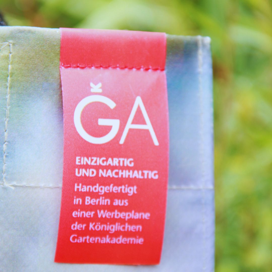 Eine Nahaufnahme des roten Stoffetiketts auf der Upcycling Tasche 'Leaf' von Werbeplane - Königliche Gartenakademie. Auf dem Etikett steht weißer deutscher Text: GA. Einzigartig und nachhaltig. Handgefertigt in Berlin. Marke: Königliche Gartenakademie.