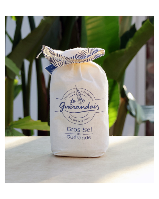 Eine Stofftasche mit der Aufschrift Le Guérandais Guerandais | Grobes Meersalz aus der Bretagne ruht auf einer hellen Fläche, hinter der sich grüne Pflanzen befinden. Der blau-weiße Kordelzug deutet auf seine authentische bretonische Herkunft hin.