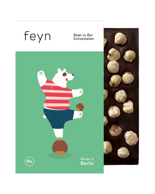 Eine Feyn-Schokoladentafel mit ganzen Bio-Haselnüssen ist teilweise in eine farbenfrohe Verpackung mit einem Cartoon-Bären in einem rot gestreiften Hemd eingewickelt. Der Text lautet "Feyn | Dunkle Schokolade mit Haselnuss" und "Made in Berlin".