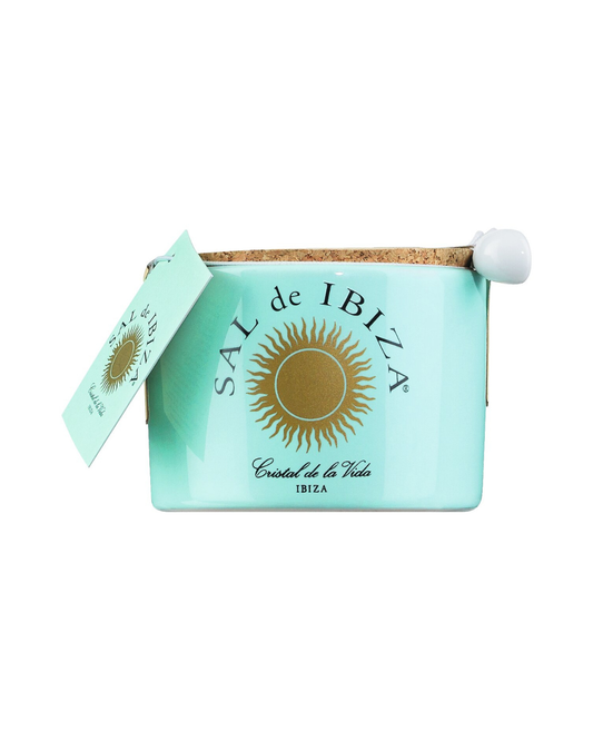 Ein hellblaues Sal de Ibiza | „Fleur de Sel“-Meersalz im 150-g-Keramikglas mit dem goldenen Sal de Ibiza-Sonnenlogo, einem Korkdeckel, einem angebrachten Etikett und einem kleinen weißen Keramiklöffel – ideal zum Servieren dieses erlesenen Meersalzes.