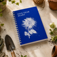 Ein blaues „KGA Gartentagebuch | ‚Five Years Diary‘“ in limitierter Auflage der Königlichen Gartenakademie mit einer Illustration einer weißen Blume liegt auf einem Gartentisch inmitten von Gartenhandschuhen, einer Gartenschaufel, Schnur, Topfpflanzen und verstreuter Erde.