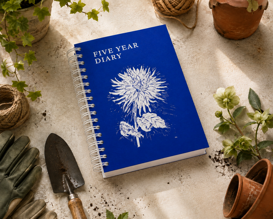 Ein blaues, spiralgebundenes KGA-Gartentagebuch „Five Years Diary“ | Limitierte Auflage der Königlichen Gartenakademie mit einer weißen Blumenillustration liegt auf einem Gartentisch, umgeben von Handschuhen, einer Gartenschaufel, Schnur, Topfpflanzen und verstreuter Erde.