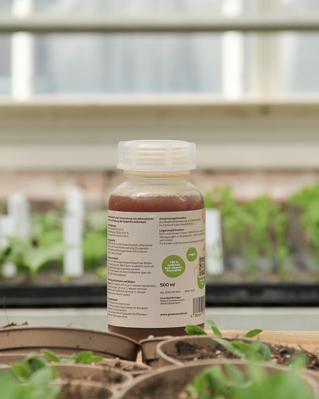 Eine 500-ml-Flasche Green Mundo | 'Bakti' Kompost- und Bodenaktivator mit weißem Verschluss und grünem Etikett steht zwischen Pflanzentöpfen und Setzlingen; die braune Flüssigkeit im Inneren ist sichtbar, ein Gewächshausfenster verschwimmt im Hintergrund.