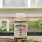 Eine 500-ml-Plastikflasche von Green Mundo | 'Bakti' Kompost- und Bodenaktivator steht auf einem Gartentisch zwischen grünen Setzlingen, mit verschwommenen Gewächshausregalen im Hintergrund.