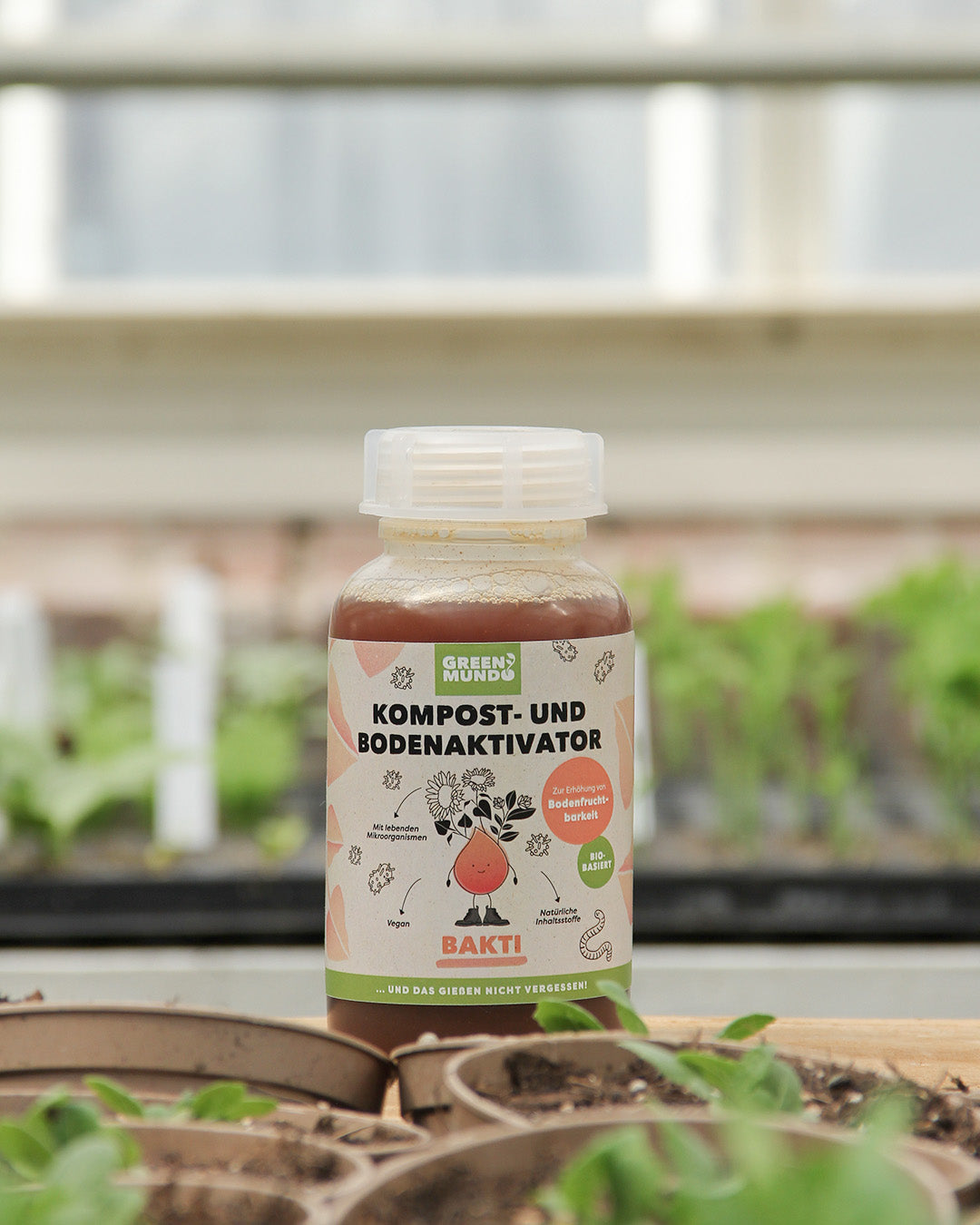 Eine 500-ml-Plastikflasche von Green Mundo | 'Bakti' Kompost- und Bodenaktivator steht auf einem Gartentisch zwischen grünen Setzlingen, mit verschwommenen Gewächshausregalen im Hintergrund.