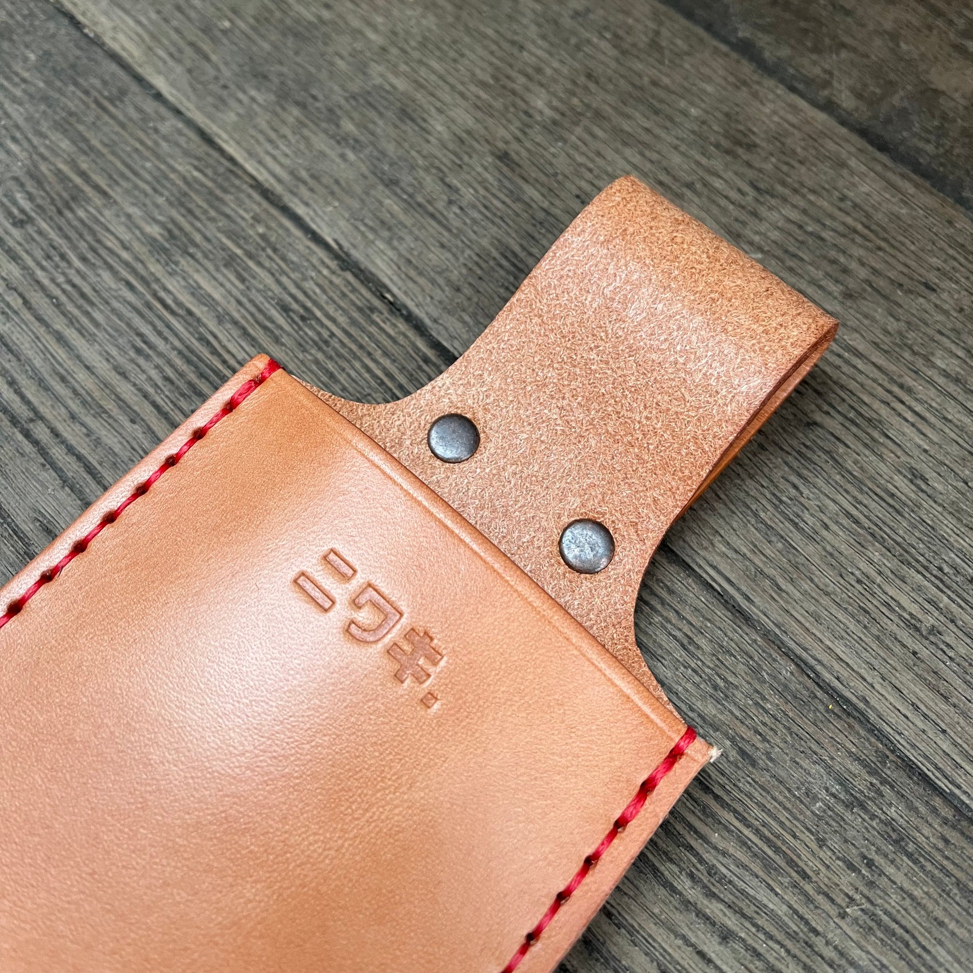 Eine Nahaufnahme der Niwaki | 'Saw Holster' hochwertigen Lederschutzhülle Klappsäge von Niwaki zeigt rote Nähte, zwei Metallnieten und geprägte japanische Schriftzeichen auf dem hellbraunen Leder, das auf einer Holzoberfläche ruht.