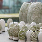 Eine Reihe kleiner Des Pots | Vase Ei-auf-Schale Form (H 10 cm, Ø 5,5 cm) der Königlichen Gartenakademie - einige schlicht weiß, andere mit grüner und weißer Blumenglasur - sind ausgestellt, dahinter zwei größere gemusterte Vasen.