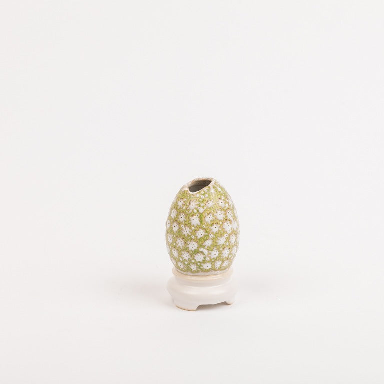 Die Des Pots | Vase Ei-auf-Schale Form klein (H 10 cm, Ø 5,5 cm) von Königliche Gartenakademie ist eine eiförmige, handgemachte Keramikvase mit grün-weißem Blumenmuster, glasiert und auf einer kleinen weißen Schale platziert.