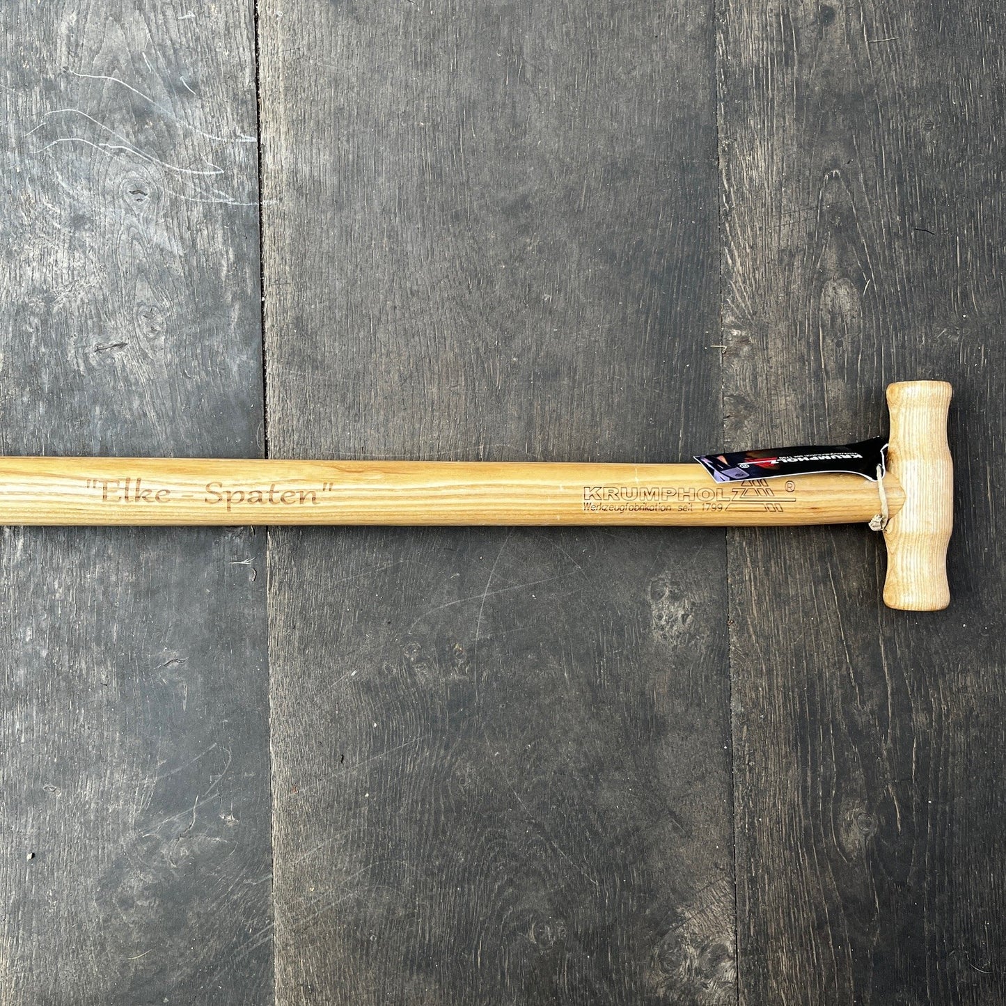 Der Krumpholz Damenspaten 'Elke' 85cm 1589 mit Holzgriff liegt auf dunklem Boden; sein leichtes, ergonomisches Design eignet sich ideal für vielseitige Gartenarbeiten. Eine schwarze und rote Markierung hängt beim Griff.