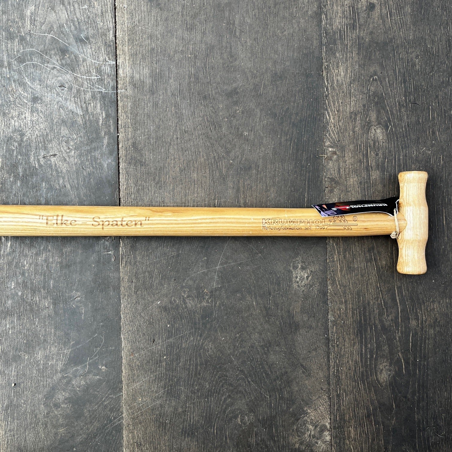 Der Krumpholz Damenspaten 'Elke' 85cm 1589 mit Holzgriff liegt auf dunklem Boden; sein leichtes, ergonomisches Design eignet sich ideal für vielseitige Gartenarbeiten. Eine schwarze und rote Markierung hängt beim Griff.