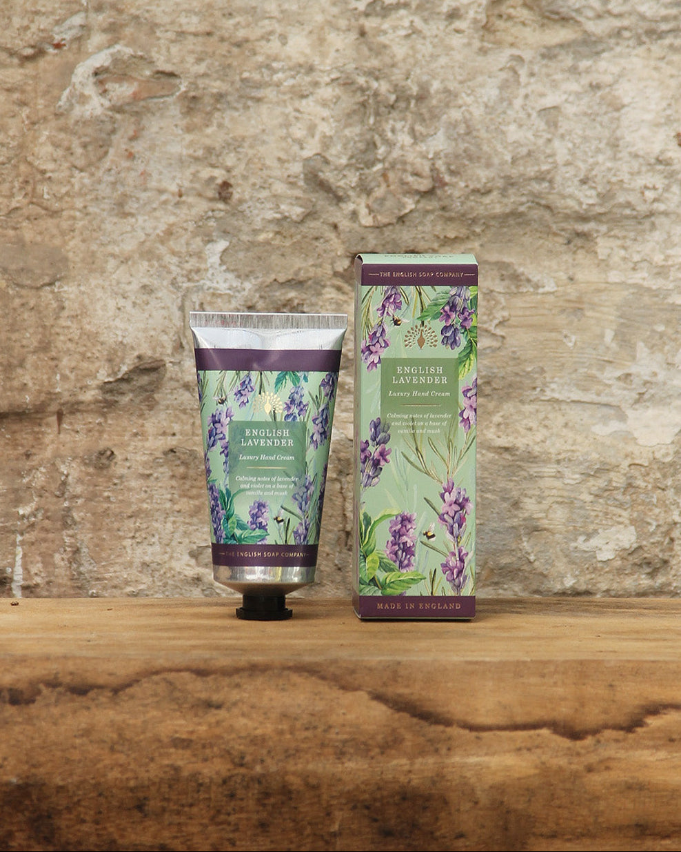 Eine Tube und eine Schachtel von The English Soap Company ESC | 'English Lavender' Handcreme 75 ml, einer luxuriösen veganen Handcreme mit floraler Verpackung, liegen auf einer Holzfläche vor einer rustikalen Steinmauer.