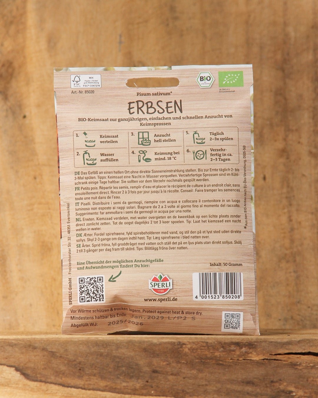 Eine Packung Sperli's Bio Keimsprossen Saatgut 'Erbsen' von Sperli's steht aufrecht auf Holz und zeigt auf der Rückseite deutsche Pflanz- und Pflegehinweise für den Anbau von Erbsensprossen.