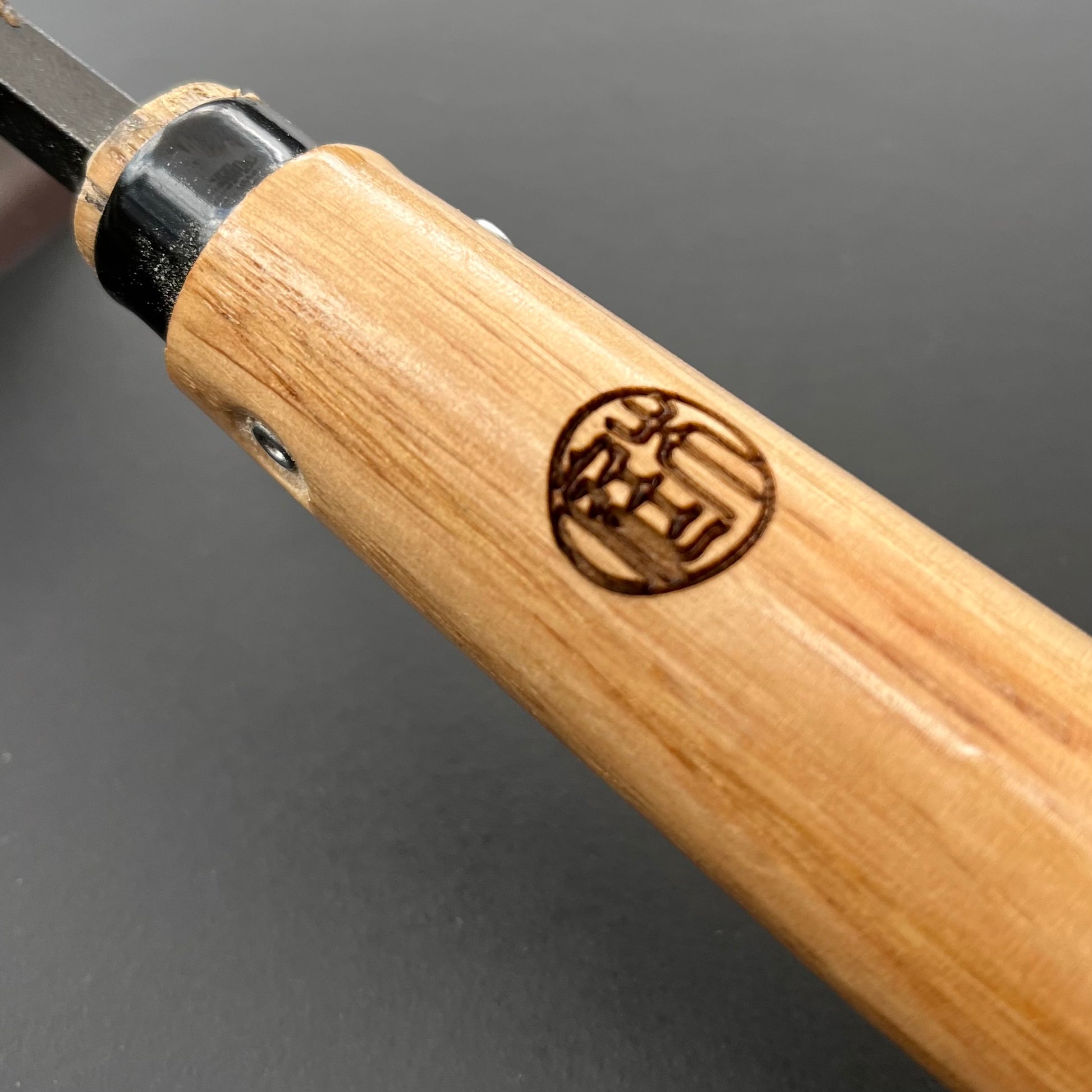 Eine Nahaufnahme zeigt den Holzgriff mit dem japanischen Logo auf der Niwaki | 'Unkrauthacke' Jäthacke für Rechts- und Linkshänder von Niwaki, der an der Metallklinge für effizientes Unkrautjäten im Garten befestigt ist, vor einem dunklen Hintergrund.