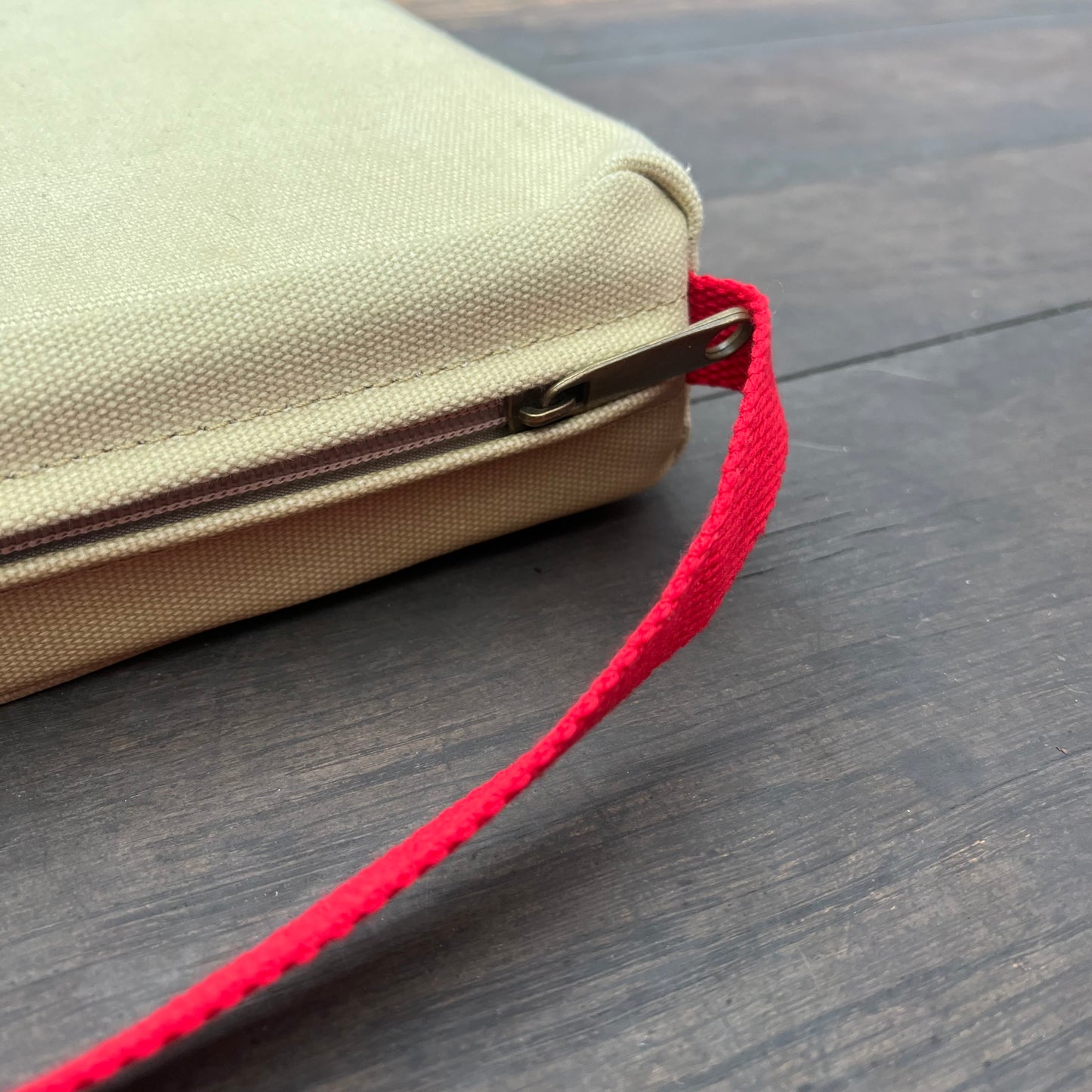 Nahaufnahme eines beigen Stoffbeutels mit Reißverschluss und roter Handschlaufe, der auf dunklem Holz ruht. Die rechteckige Tasche mit dem teilweise sichtbaren Reißverschluss ist ideal für den Transport von wichtigen Dingen wie dem Niwaki | Kniekissen von Niwaki.