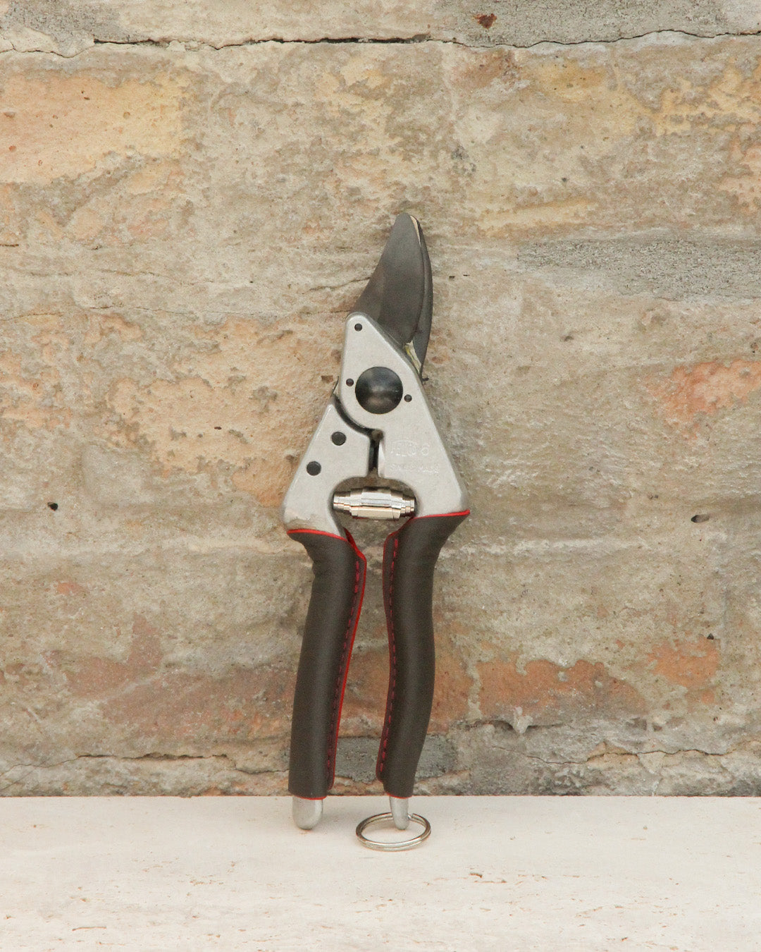 Ein Paar der schwarz-silbernen Felco '6 Elite' Kompakte Gartenschere mit rot abgesteppten Griffen und ergonomischem Design steht aufrecht vor einem strukturierten Steinmauer-Hintergrund.