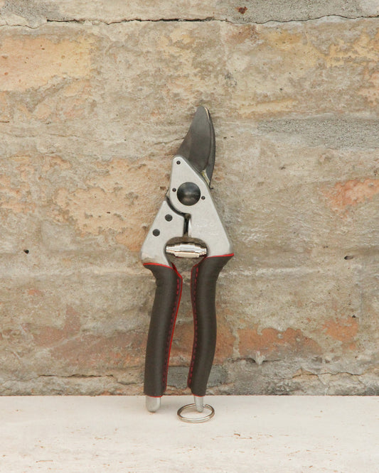 Ein Paar der schwarz-silbernen Felco '6 Elite' Kompakte Gartenschere mit rot abgesteppten Griffen und ergonomischem Design steht aufrecht vor einem strukturierten Steinmauer-Hintergrund.
