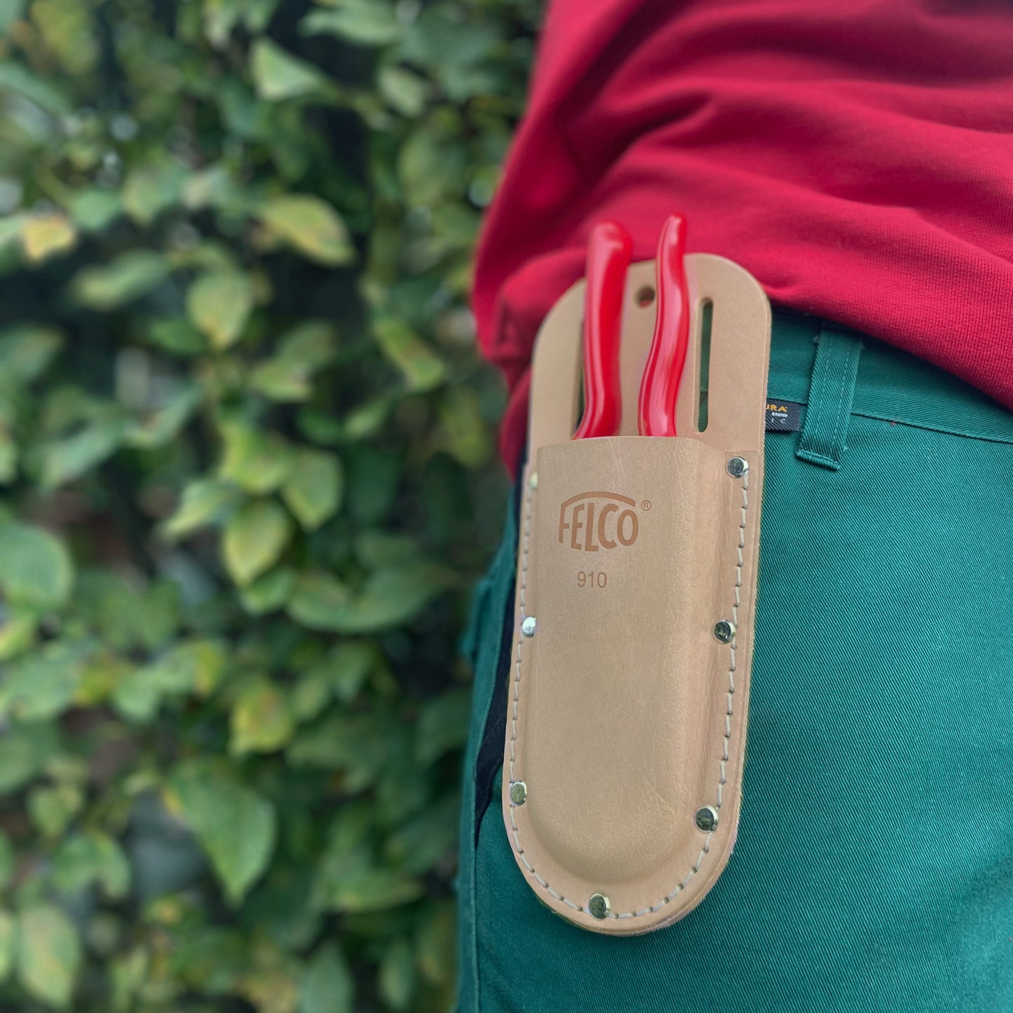 Eine Person in grüner Hose und rotem Hemd trägt ein Felco 910 Echtleder Scherenetui von Felco mit Gartengeräten mit rotem Griff am Gürtel. Im Hintergrund ist unscharfes grünes Laub zu sehen.