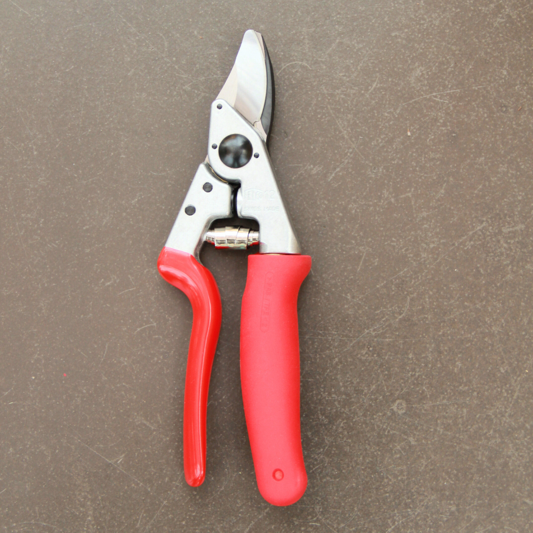 Ein Paar Felco 12 Gartenscheren von Felco mit roten Griffen und einer silbernen Klinge ruht auf einer braunen, strukturierten Oberfläche - perfekt für Präzisionsschnitte.