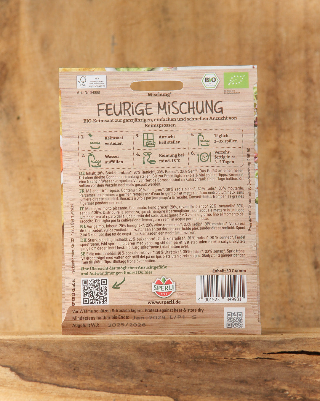 Rückseite einer Sperli's Bio Keimsprossen Saatgut 'Feurige Mischung' Packung mit deutscher Anleitung für den Anbau von würzigen Keimsprossen, mit Logos, Barcode, und auf einer Holzunterlage.