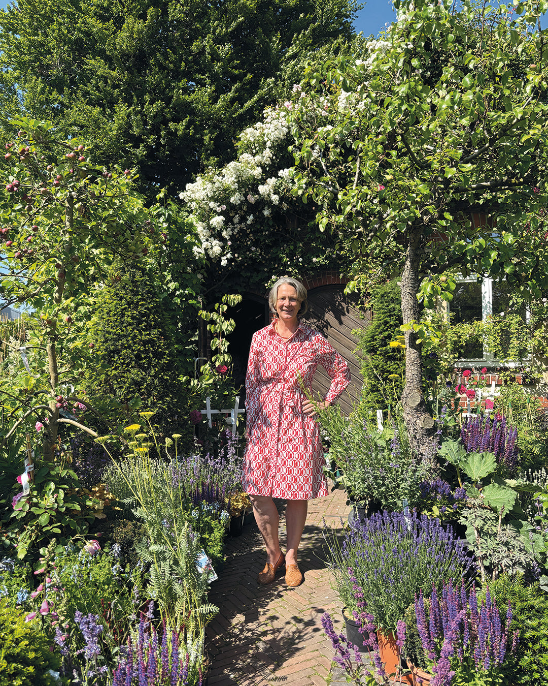 Eine Frau in einem rot-weißen Kleid lächelt auf einem sonnigen Gartenweg - ein inspirierender Anblick für alle Garteninteressierten beim HALBTAGESKURS "Der Garten als Lebensraum NEU" der Königlichen Gartenakademie am Mi. 18.02.2026, 09.30-12.30.