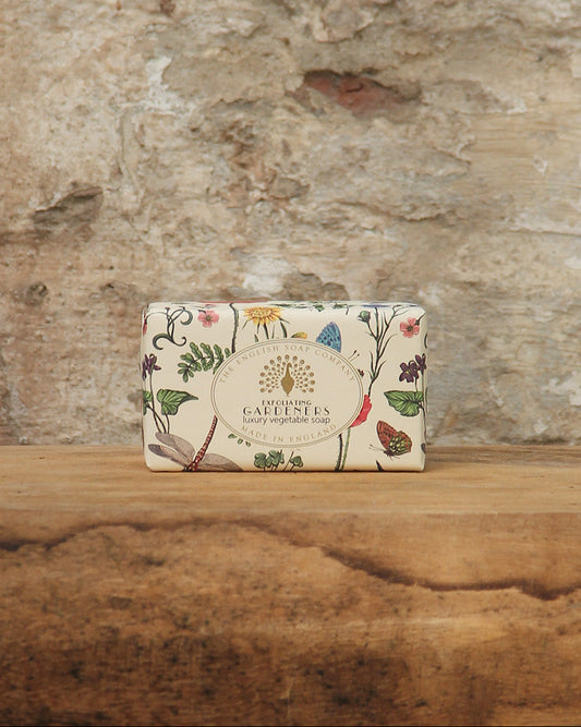 Eine Tafel ESC | 'Gardeners' fruchtig duftende pflanzliche Handseife von The English Soap Company, eingewickelt in blumengemustertes Papier, ruht auf Holz vor einer verwitterten Steinwand.