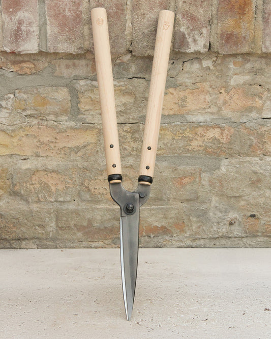 Ein Paar Niwaki | 'Niwaki Garden Shears' Heckenschere von Niwaki mit leichten FSC-zertifizierten Holzgriffen und scharfen Metallklingen steht aufrecht vor einer rustikalen Ziegelwand und einer Betonoberfläche.