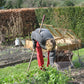 Eine Person in einem grauen Pullover und einer roten Hose, die mit einer Hacke im Garten Frühjahrsarbeiten verrichtet, inspiriert von Sa., 13.3.2027 | 11.00 – 12.30 Uhr GARTENTALK – Gedanken zum Gärtnern im Frühling.